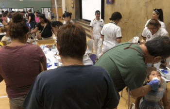 Health professionals provide dental care to children and adults at a busy indoor community event with multiple tables and attendees.