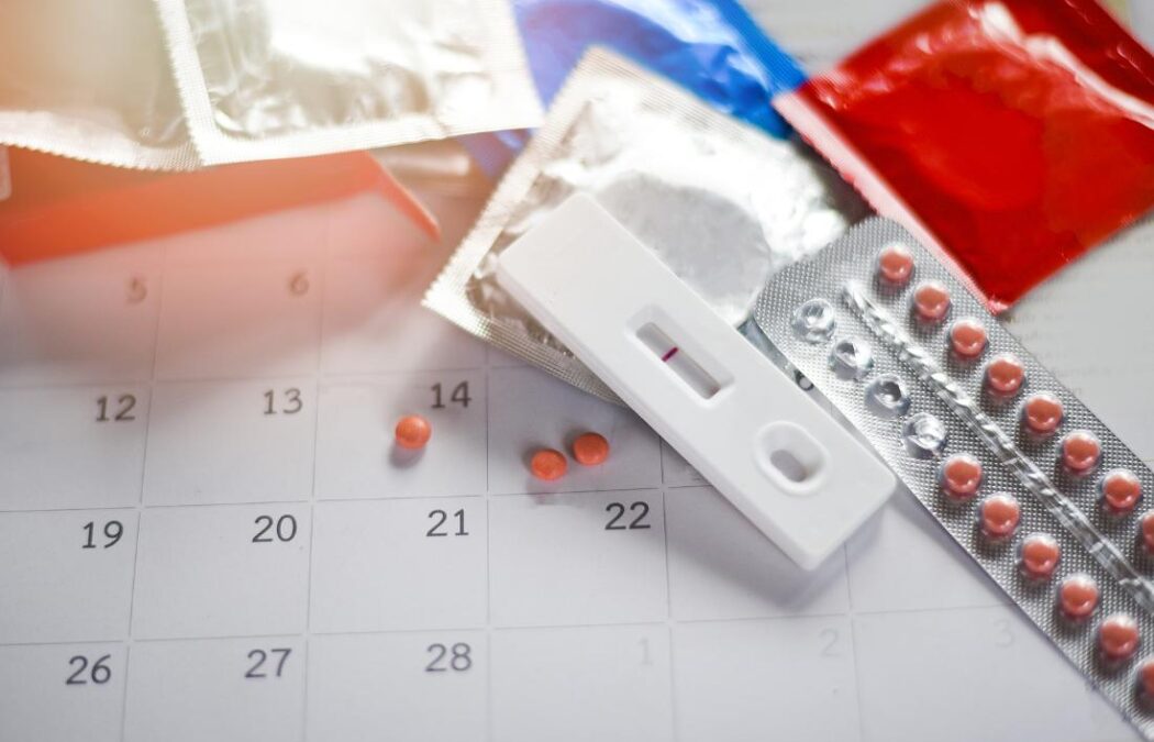 A pregnancy test, pill blister pack, pills, and colored condom wrappers are on a calendar.