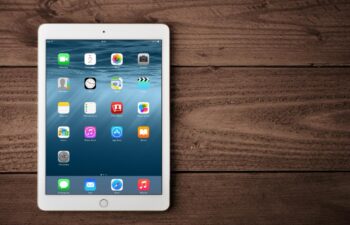 A white iPad with the home screen displayed is placed on a wooden surface.