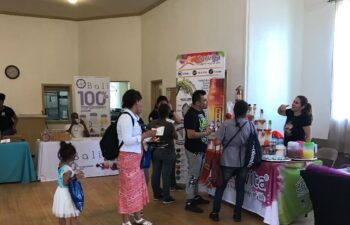 People visit vendor tables at an indoor community event. A woman hands samples to attendees, while others browse displayed products. Natural light comes from a window on the right.