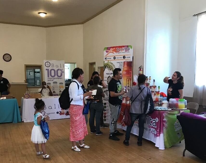People visit vendor tables at an indoor community event. A woman hands samples to attendees, while others browse displayed products. Natural light comes from a window on the right.