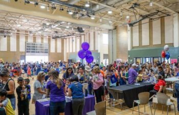 Large group of people gathered in a gymnasium for a community event with tables, informational booths, and balloons.