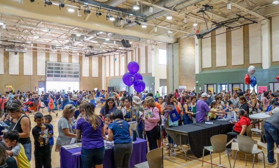 Large group of people gathered in a gymnasium for a community event with tables, informational booths, and balloons.