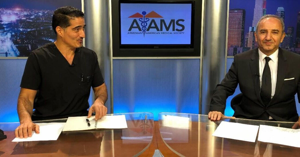 Two men sit at a news desk with papers. One wears black scrubs, the other a suit. The screen behind them displays the Armenian American Medical Society logo.