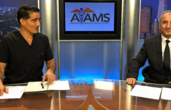 Two men sit at a news desk with papers. One wears black scrubs, the other a suit. The screen behind them displays the Armenian American Medical Society logo.