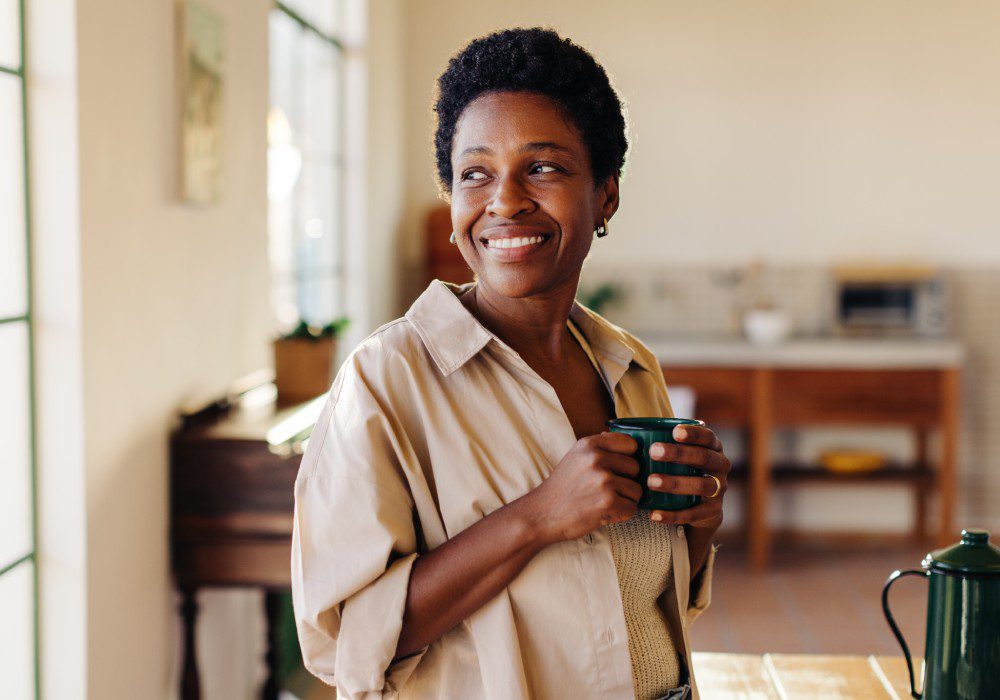 Happy Brazilian woman looking outside the window in her home kitchen, smiling and holding an enamel mug of plain coffee.