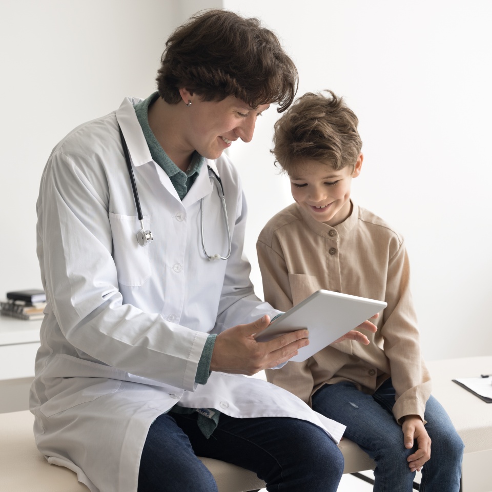 Let me explain. Happy little child sit on medical couch at pediatric cabinet close to friendly confident male doctor look on tablet screen listen about importance of vaccines learn to protect health