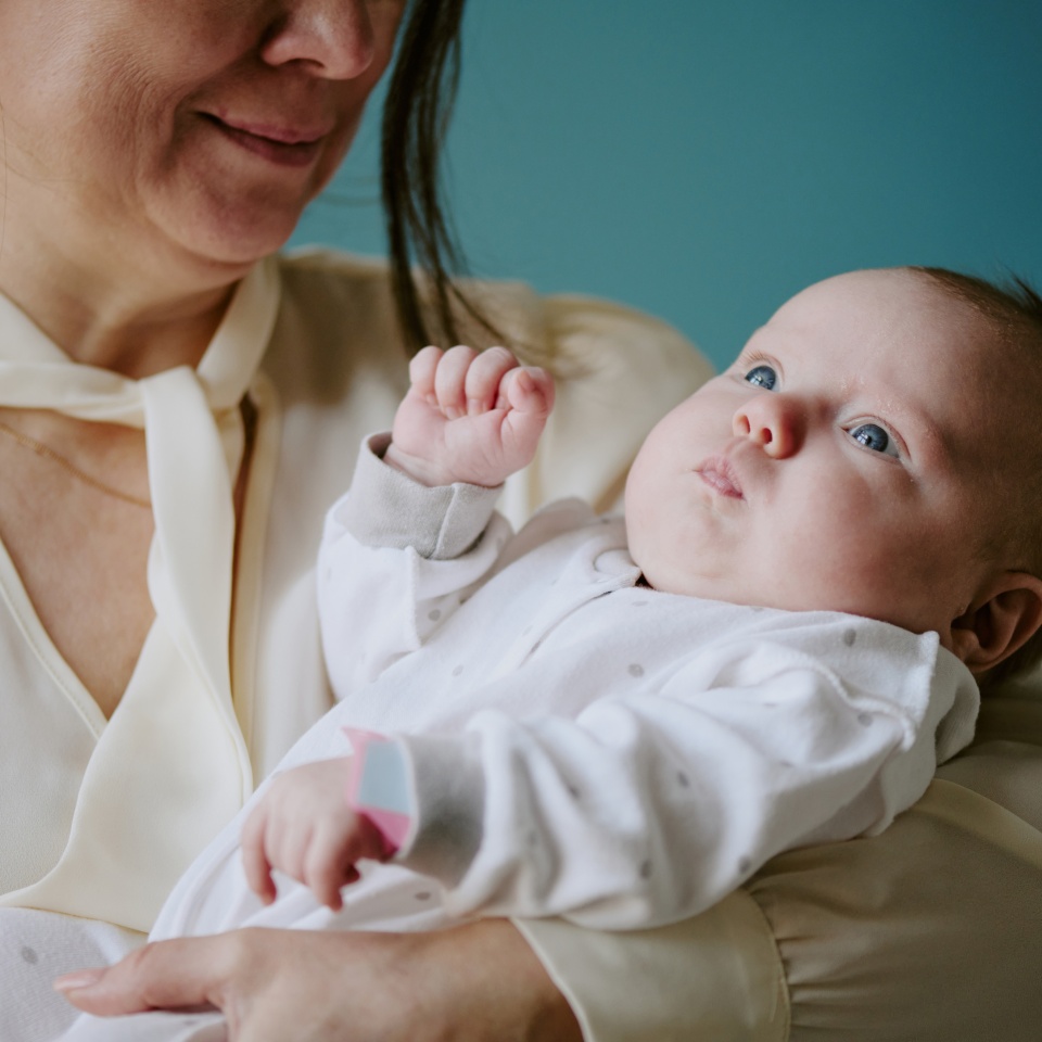 Woman holding newborn baby in vibrant beige room. Baby wearing white onesie, gazing upwards while mother lovingly holding infant close