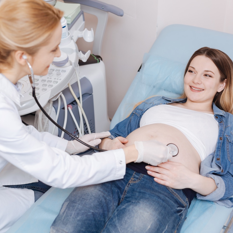 Examining fetus development . Friendly professional female obstetrician working in the sonography cabinet while examining pregnant belly and using stethoscope for detecting heat beating