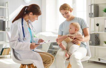 Mother and child visit pediatrician at clinic. Happy young woman holding her cute little baby on her lap while sitting on chair and talking to friendly, smiling doctor. Children's healthcare concept