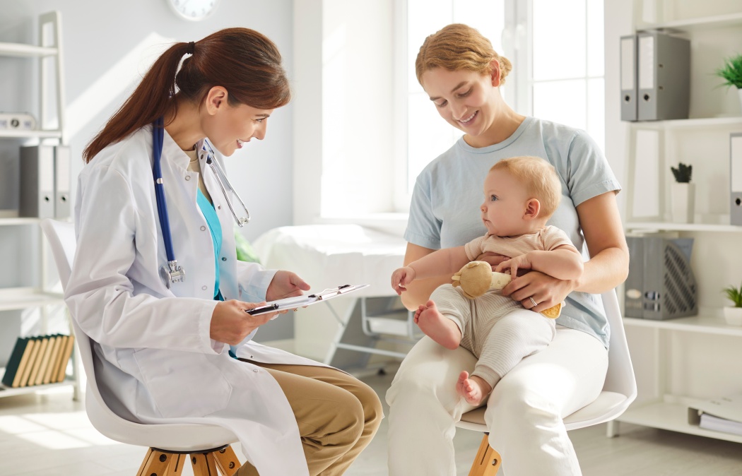Mother and child visit pediatrician at clinic. Happy young woman holding her cute little baby on her lap while sitting on chair and talking to friendly, smiling doctor. Children's healthcare concept