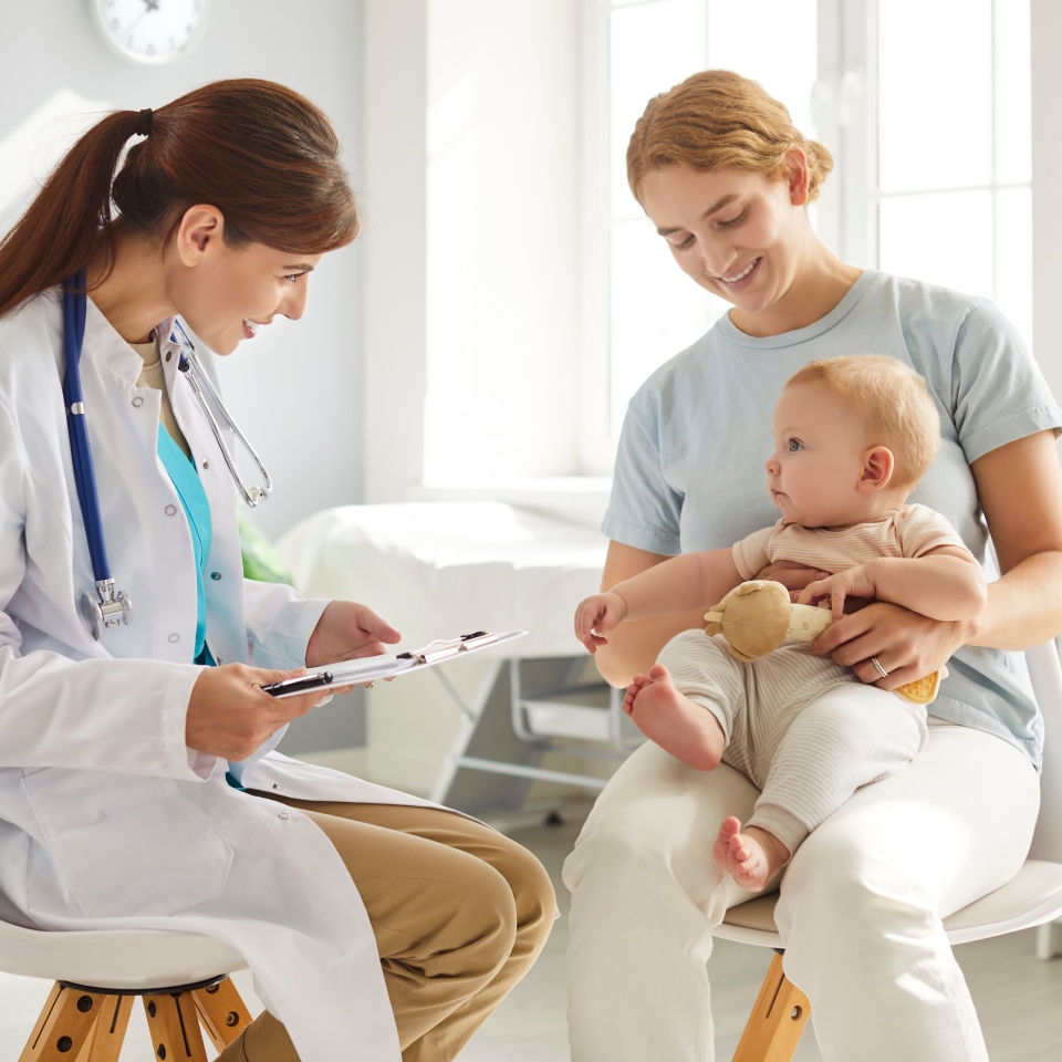Mother and child visit pediatrician at clinic. Happy young woman holding her cute little baby on her lap while sitting on chair and talking to friendly, smiling doctor. Children's healthcare concept