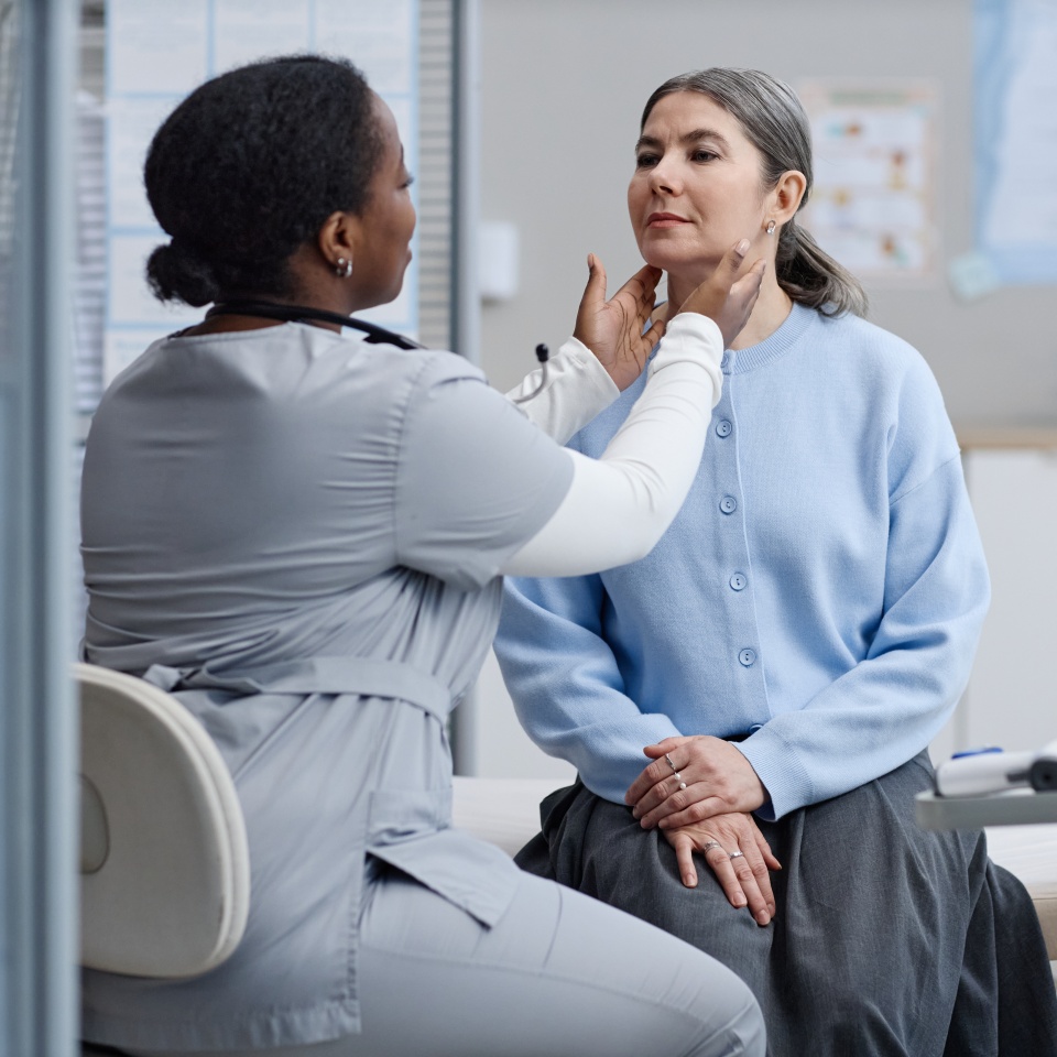 Black woman doctor examining neck of middle aged Caucasian woman during medical checkup in clinic, both women focused on health assessment, medical equipment visible on table nearby