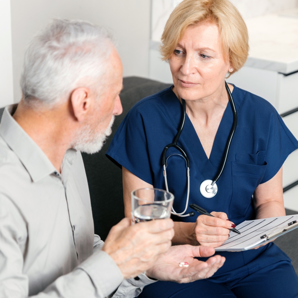 Doctor is discussing medication options and treatment plan with elderly patient at home. Caregiver make records in medical card. Man holds glass of water and pills, listening benefits and side effects