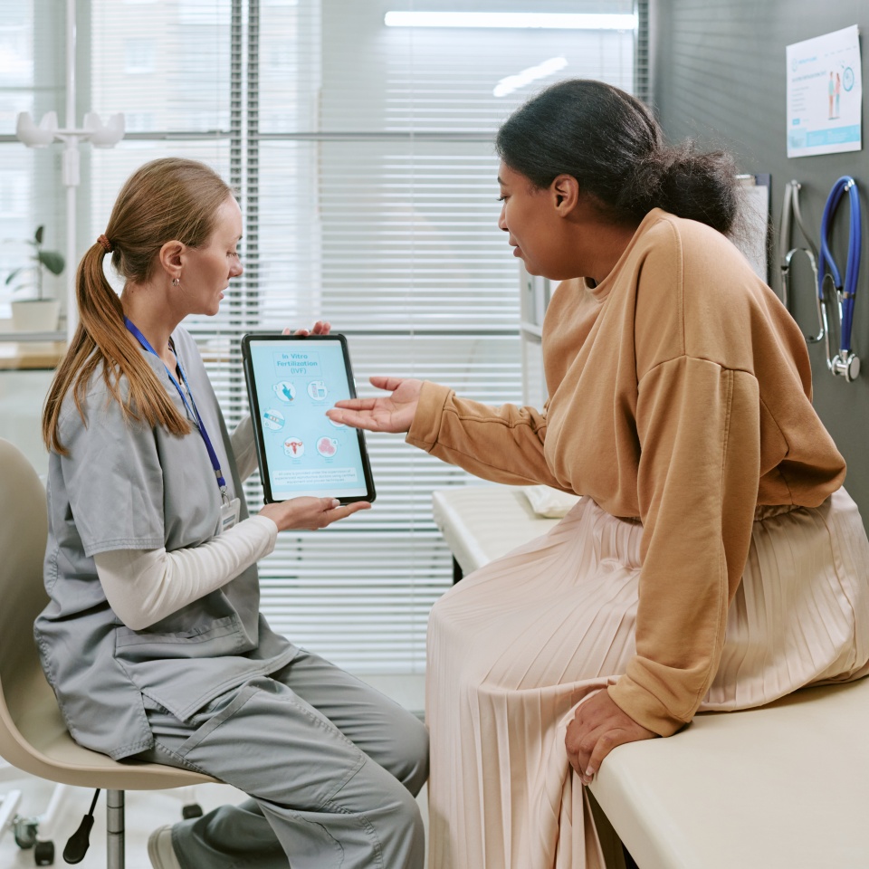 Black woman consulting female doctor, discussing HPV information on tablet during clinic visit. Supporting gynecology education, preventive care marketing, medium shot