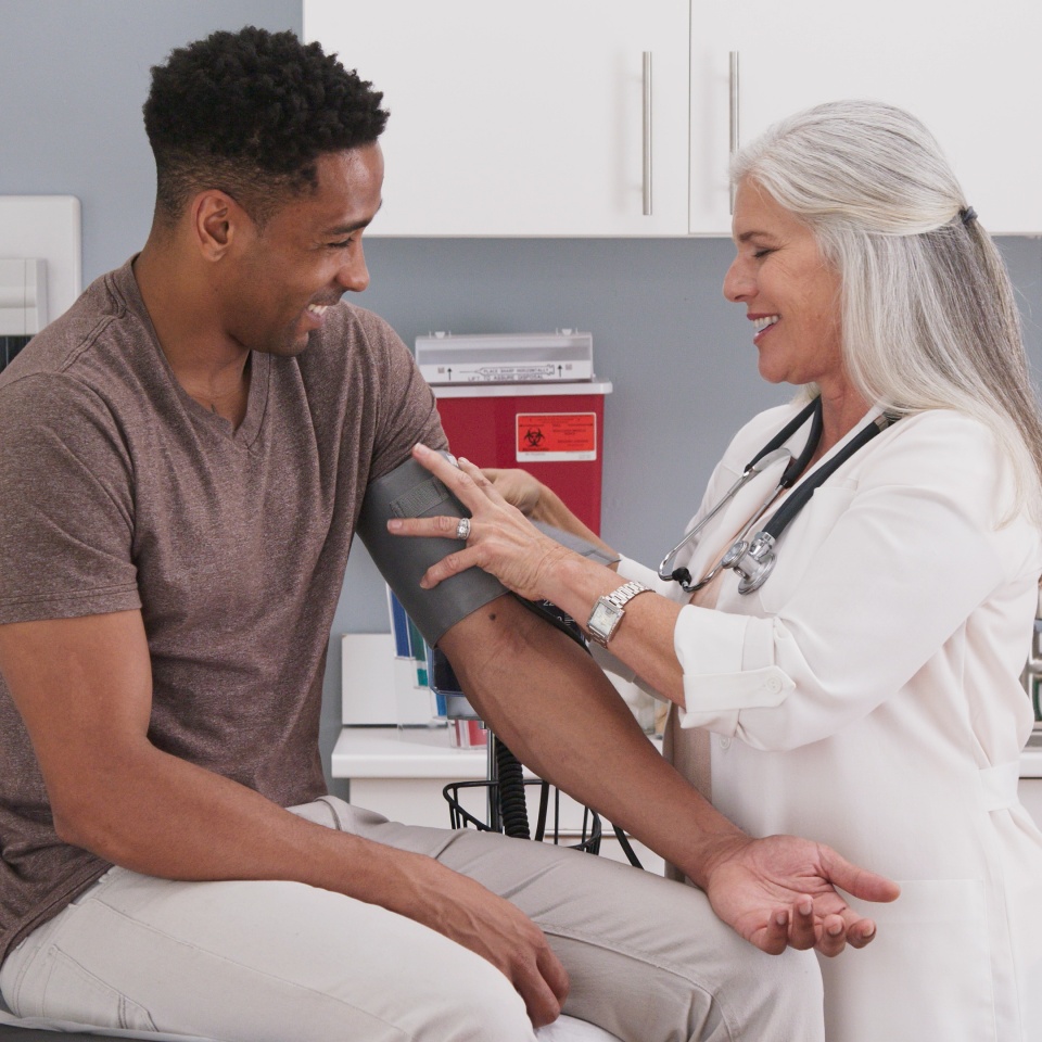 A doctor checks a patient’s blood pressure in a medical office. The patient sits on an exam table and smiles as the doctor secures the cuff on his arm.