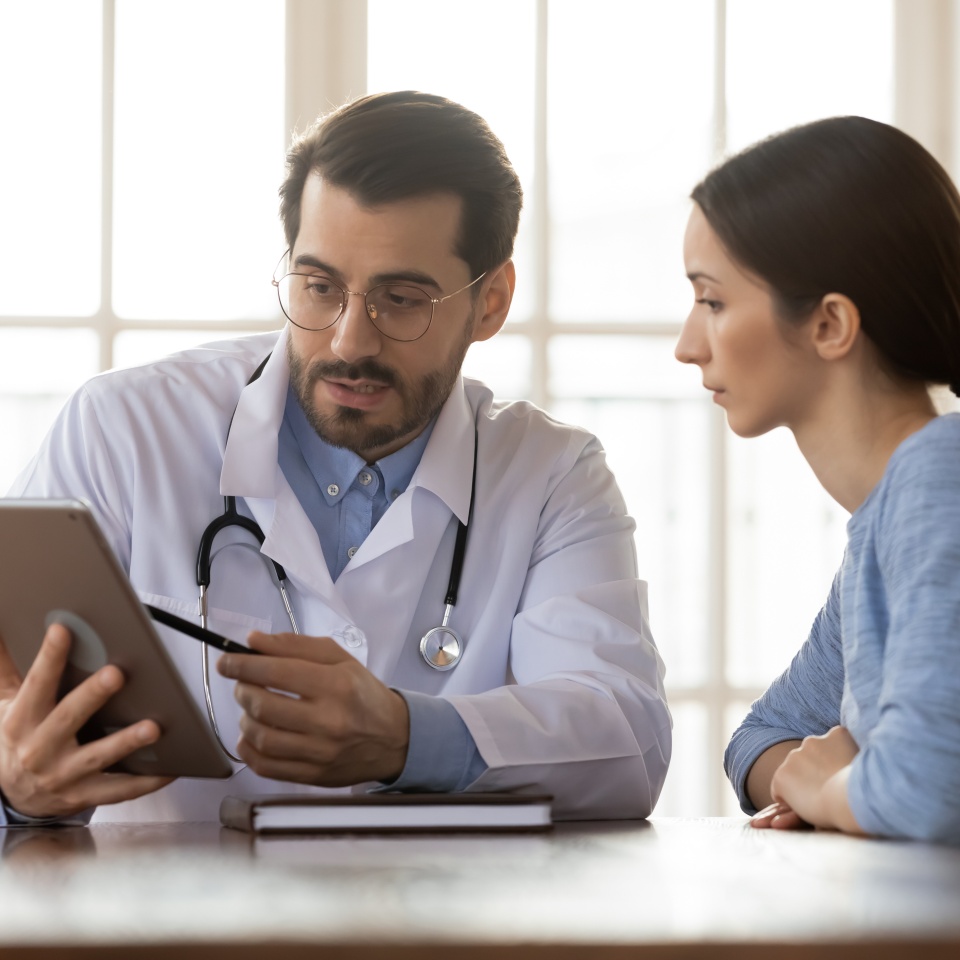 Focused young male doctor pointing at digital computer tablet screen, sharing health tests laboratory results to serious female patient or explaining medical insurance benefits at meeting in clinic.