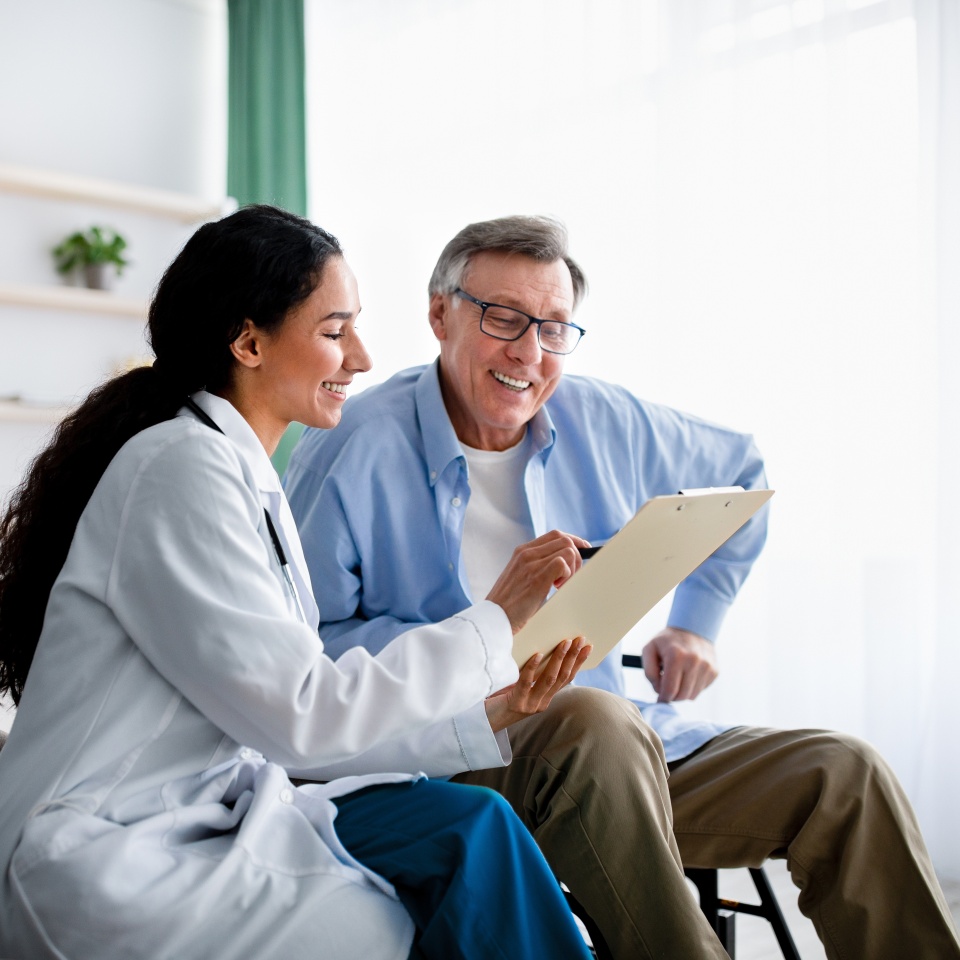 Young doctor asking senior impaired male patient in wheelchair to sign insurance policy at home. Handicapped elderly man putting his signature under surgery consent form, reading medical document