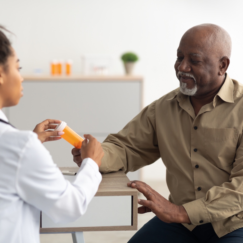 Pharmaceutical Drug Prescription Concept. Young black doctor showing elderly male patient pills bottle, pointing at tablets. Female specialist prescribing mature man treatment, painkiller or vitamin