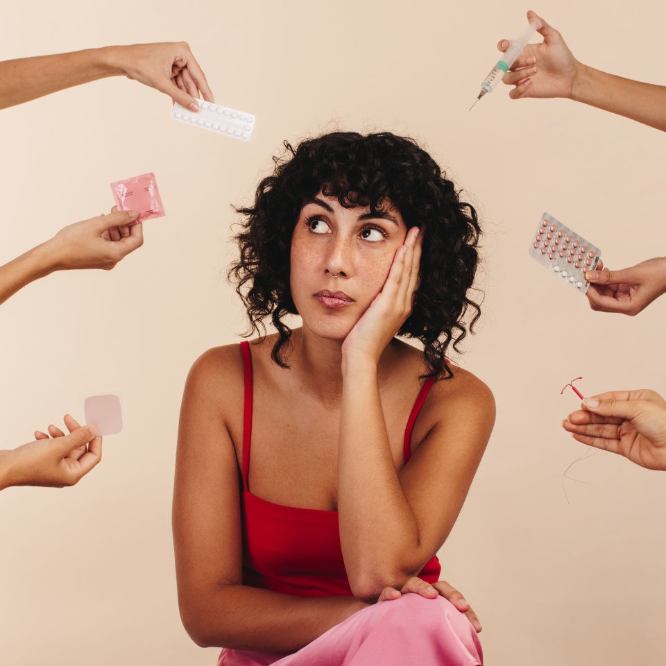 Young woman looking away thoughtfully while surrounded by hands holding different forms of hormonal and non-hormonal contraception. Pensive young woman making a decision about her reproductive health.
