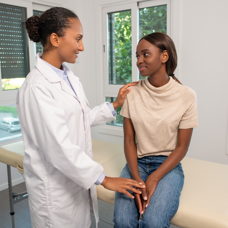 Smiling general practitioner comforting young female patient in clinic. Young African American woman sitting on coach and talking to doctor in hospital. Visiting doctor concept
