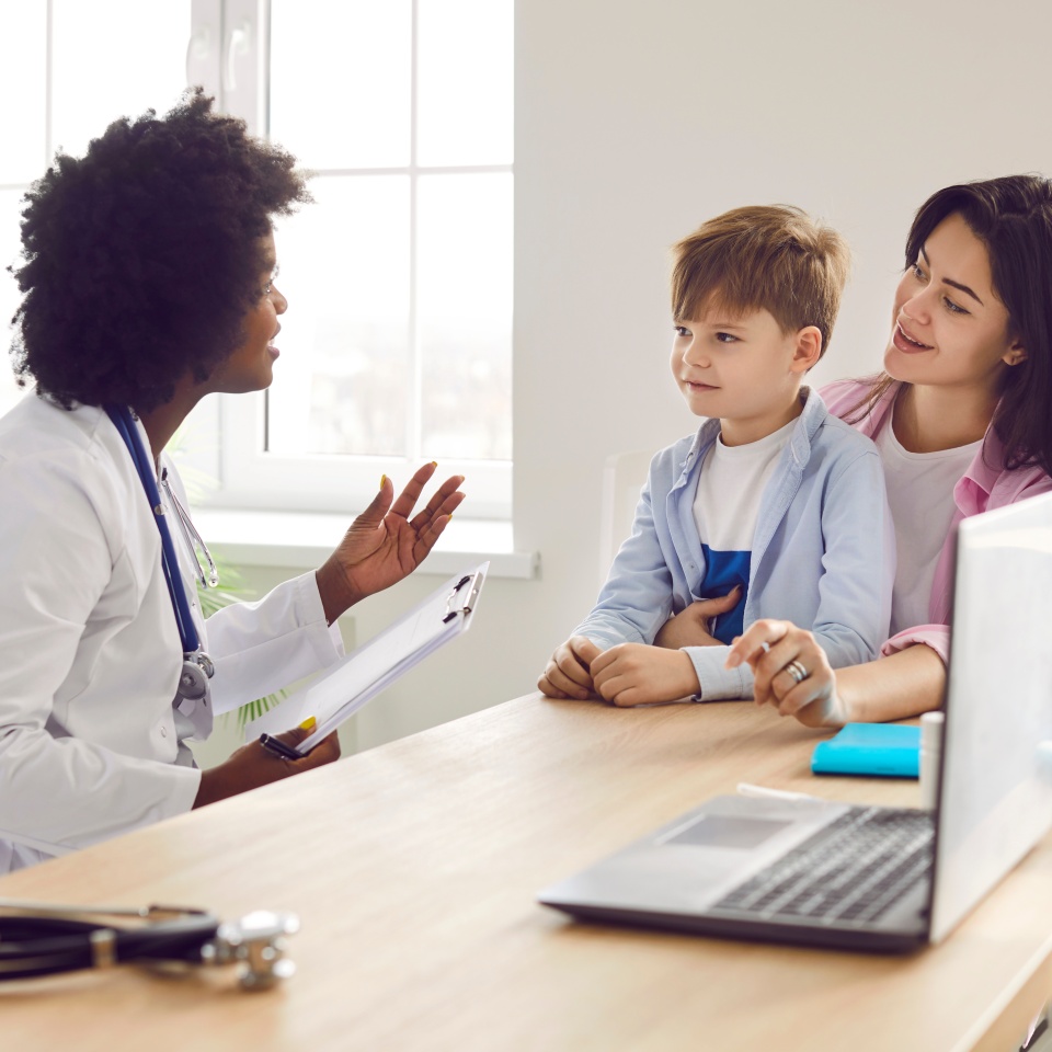 Little boy patient with his mother talking with friendly african american pediatrician doctor on consultation in her office in clinic and listening the recommendations after medical examination.
