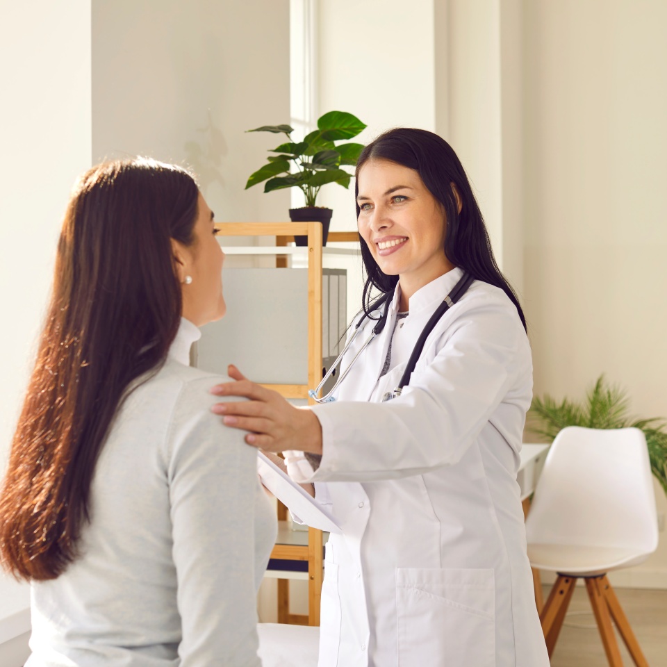 Friendly woman doctor supporting her female patient during medical examination in hospital. Smiling lady in white coat talking to young woman, calming her down and telling about effective treatment