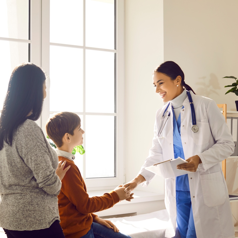 Portrait of a friendly smiling female doctor pediatrician shaking hand with a little boy patient standing with his mother and listening the recommendations after medical examination.