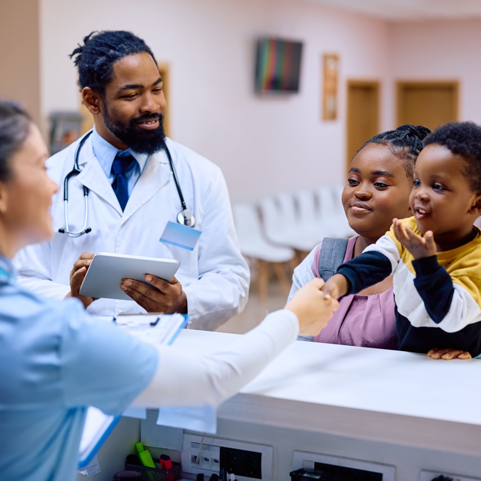 A doctor and nurse assist a woman and her young child at a clinic reception desk; the child is smiling and reaching toward the nurse.