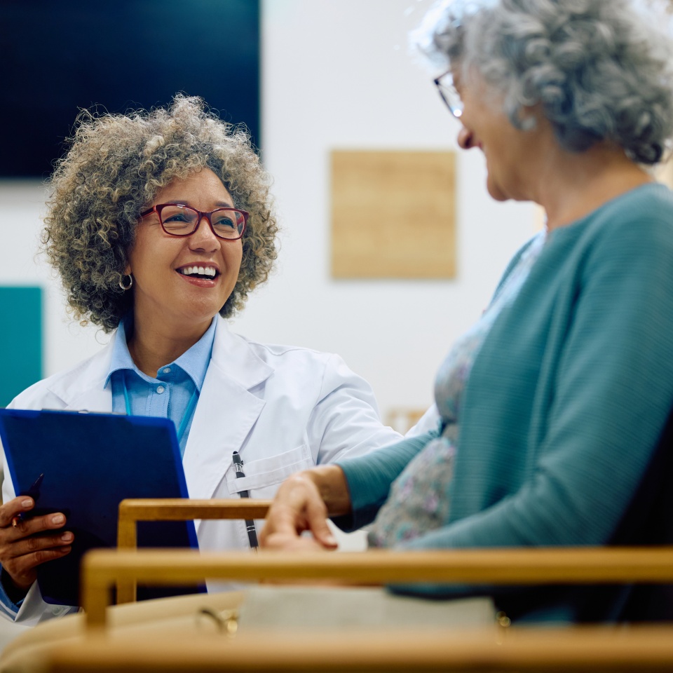 A doctor holding a clipboard smiles and talks with an older woman sitting across from her in a medical office.