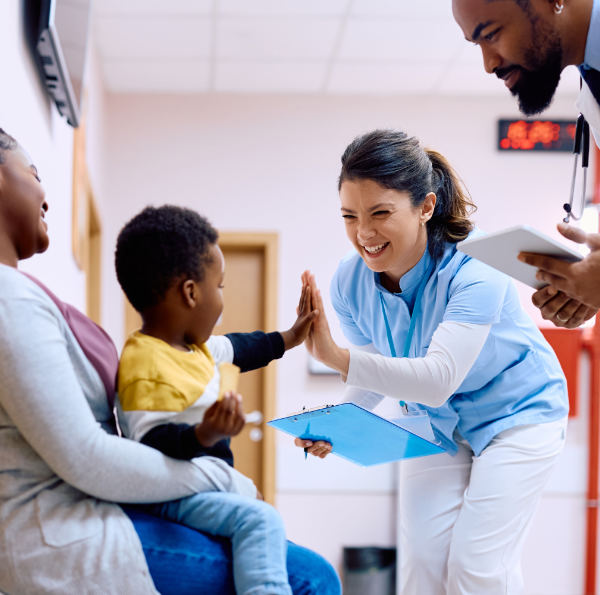 A nurse gives a high five to a young boy sitting on his mother's lap while a doctor stands nearby holding a tablet in a clinic hallway.