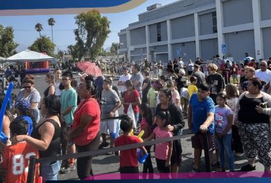 A large group of people, including adults and children, stand in line outdoors near a gray building on a sunny day.