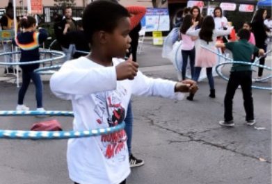A boy in a white shirt uses a blue hula hoop, with other children hula hooping in the background on an outdoor paved area.