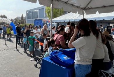 People stand in line outdoors under a tent, receiving blue giveaway bags from staff at a community event.