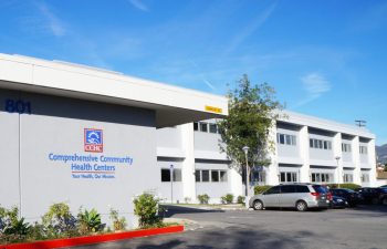A two-story white building houses the Comprehensive Community Health Centers, with parked cars in front, under a clear blue sky.