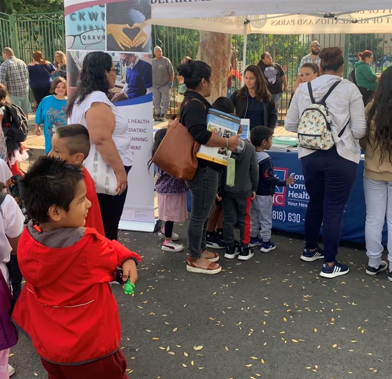 People wait in line at an outdoor community health fair booth. Children and adults stand near informational tables and banners on a sunny day.
