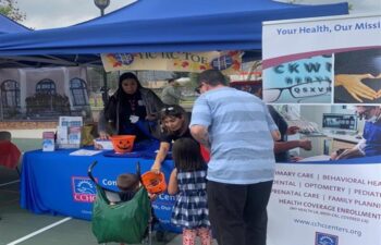 Two adults assist two children at a health information booth under blue tents during an outdoor community event. Pamphlets and trick-or-treat buckets are visible on the table.