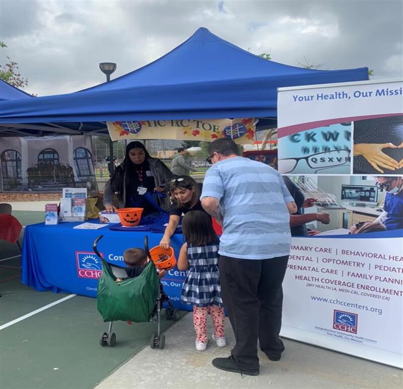 Two adults assist two children at a health information booth under blue tents during an outdoor community event. Pamphlets and trick-or-treat buckets are visible on the table.