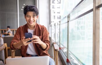 Happy Asian man using mobile phone and working with computer laptop while sitting in coffee shop