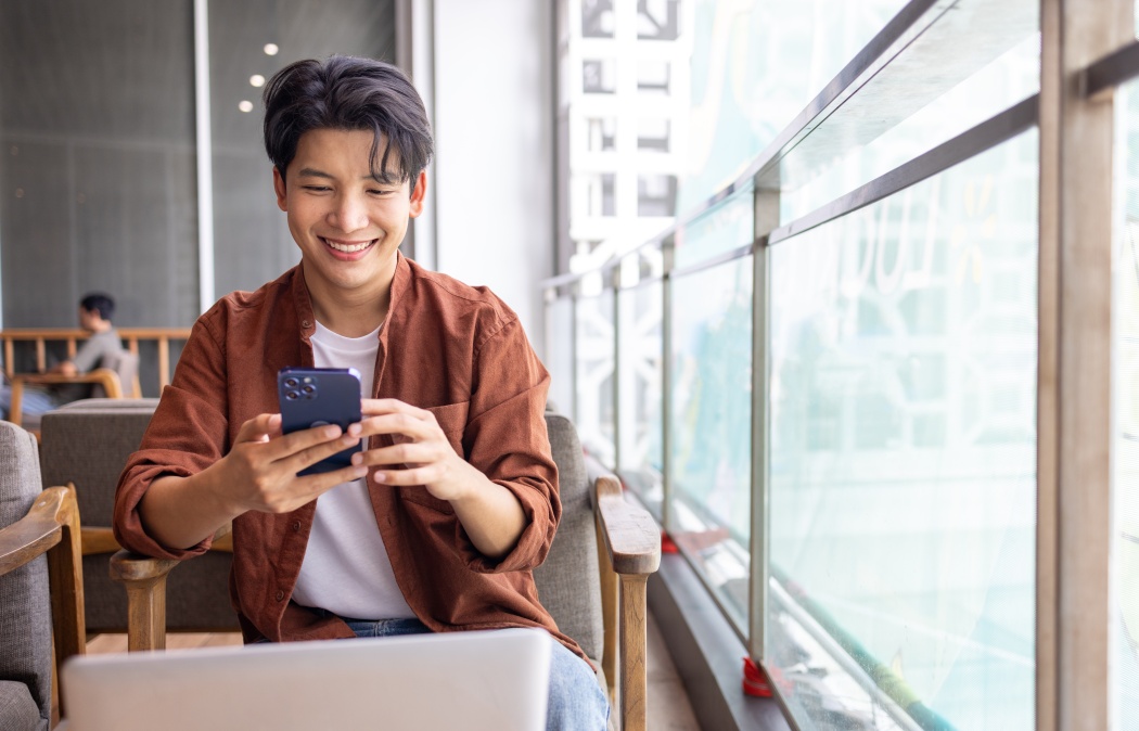 Happy Asian man using mobile phone and working with computer laptop while sitting in coffee shop