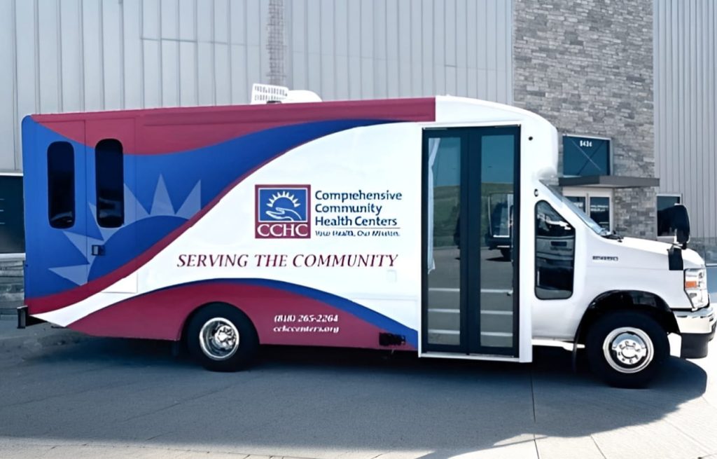 A mobile health clinic van with Comprehensive Community Health Centers branding and the phrase "Serving the Community" parked on a street.
