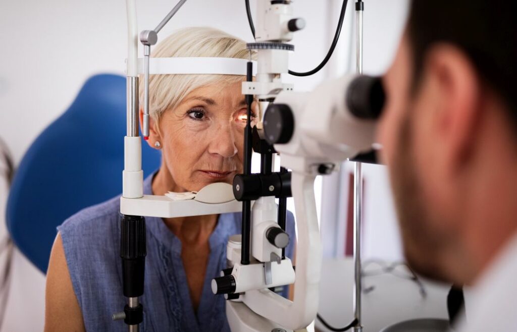 An older woman undergoes an eye examination using a slit lamp while an eye care professional observes.