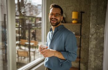 Smiling man standing by a window, enjoying a refreshing glass of water while looking outside, embracing wellness and healthy lifestyle in a comfortable indoor setting