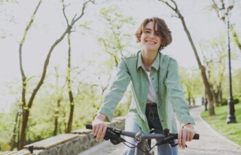 Young sporty smiling happy woman 20s wear casual green jacket jeans riding bicycle bike on sidewalk in city spring park outdoors, look camera. People active urban healthy lifestyle cycling concept.
