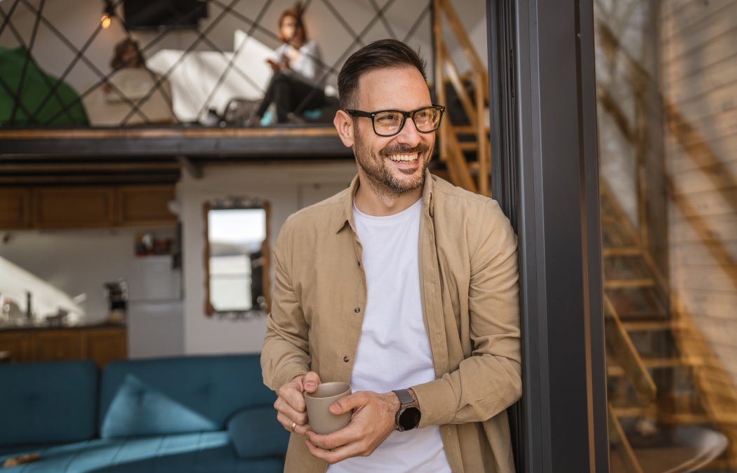 One man adult caucasian male with beard and eyeglasses stand on balcony at door in day happy smile hold cup of coffee copy space morning daily routine concept