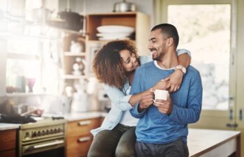 Love, coffee and happy couple in kitchen, morning hug in home with smile, hugging and healthy relationship. Happiness, man and woman embrace with affection, romance and young people in apartment