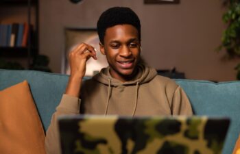 Happy black male student at cozy home watching a movie or TV show on his laptop, using the headphones to block out background noise and fully immerse himself in the content.